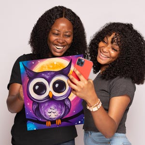 May include: Two people are smiling and holding a painting of a purple owl with big eyes in front of a yellow moon. The owl is perched on a branch with a starry background.
