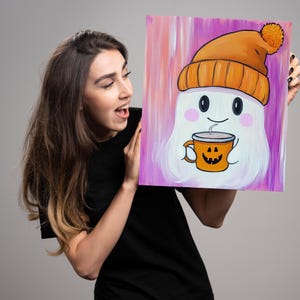 May include: A white ghost wearing an orange knit hat with a white pom pom holds a pumpkin-shaped mug with a steaming beverage. The ghost has a smiling face with rosy cheeks. The background is a purple and pink ombre.