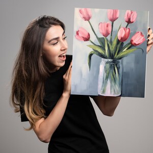 May include: A woman is holding a painting of pink tulips in a clear glass vase. The tulips are arranged in a bouquet and the vase is sitting on a light blue background.