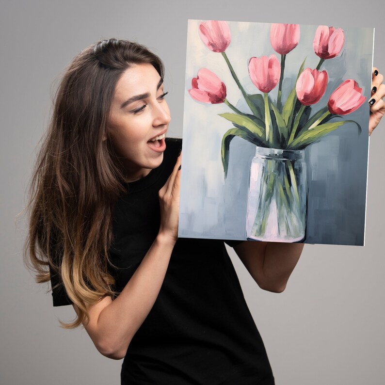 May include: A woman is holding a painting of pink tulips in a clear glass vase. The tulips are arranged in a bouquet and the vase is sitting on a light blue background.