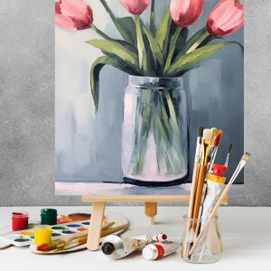 May include: A painting of a bouquet of pink tulips in a clear glass vase on an easel. The vase is sitting on a wooden surface. There are paintbrushes and paint tubes in the foreground.