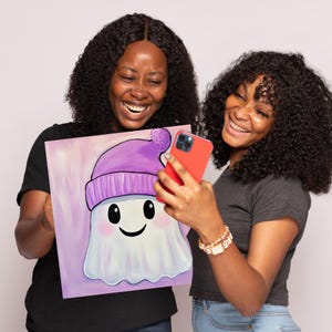 Puede incluir: Dos personas sostienen una pintura de un fantasma blanco con un sombrero morado. El fantasma sonr&iacute;e y tiene ojos grandes. La pintura tiene un fondo rosa.