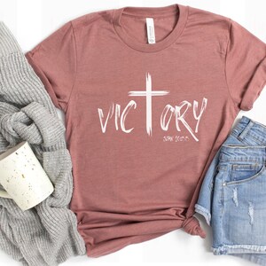 May include: A mauve t-shirt with a white cross and the word "VICTORY" in a brushstroke font. The text "JOHN 16:33" is written below the word "VICTORY".