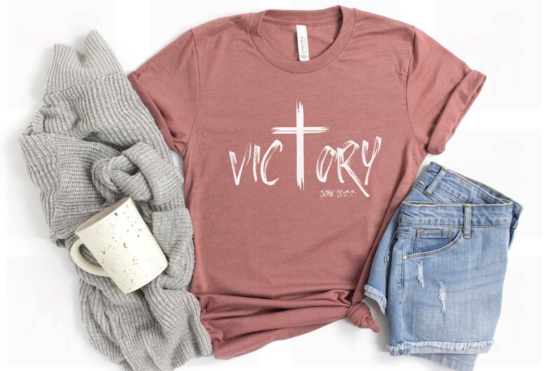 May include: A mauve t-shirt with a white cross and the word "VICTORY" in a brushstroke font. The text "JOHN 16:33" is written below the word "VICTORY".