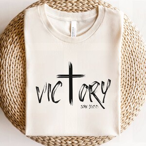May include: A white t-shirt with a black cross and the word "VICTORY" written in a brushstroke font. The text "JOHN 16:33" is written below the word "VICTORY".