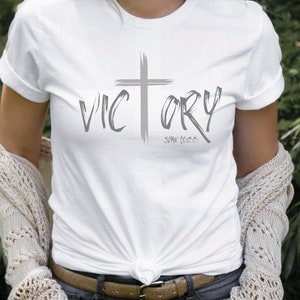 May include: White t-shirt with a gray cross and the word "VICTORY" in a brushstroke font. The text "John 16:33" is written below the word "VICTORY".