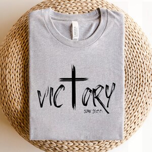 May include: A gray t-shirt with a black cross and the word "VICTORY" written in a bold, stylized font. The text "JOHN 16:33" is written below the word "VICTORY".