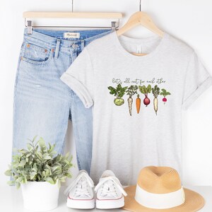 May include: A grey t-shirt with a graphic of vegetables and the text "Let's all root for each other". The shirt is styled with blue jeans, white sneakers, and a straw hat.