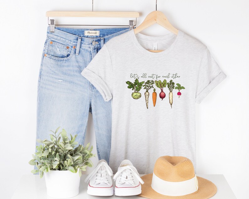 May include: A grey t-shirt with a graphic of vegetables and the text "Let's all root for each other". The shirt is styled with blue jeans, white sneakers, and a straw hat.