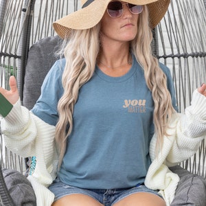 May include: A woman wearing a blue t-shirt with the text "you matter" in a light brown color. She is wearing a white cardigan and a straw hat with sunglasses.