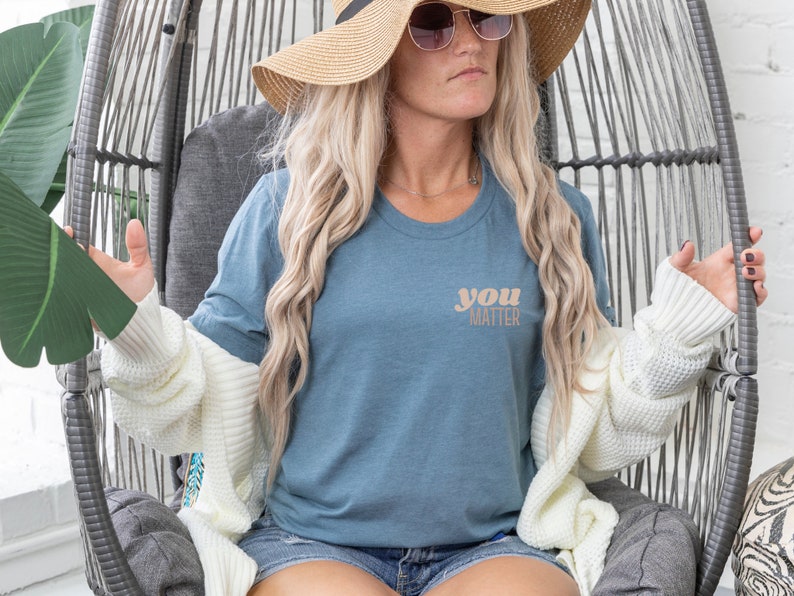 May include: A woman wearing a blue t-shirt with the text "you matter" in a light brown color. She is wearing a white cardigan and a straw hat with sunglasses.