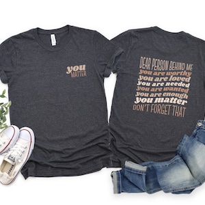 May include: A dark heather grey t-shirt with the text "you matter" on the front and the text "Dear person behind me you are worthy you are loved you are needed you are wanted you are enough you matter don't forget that" on the back.