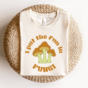 May include: A white t-shirt with a brown and orange mushroom graphic and the text "I put the fun in fungi" in a retro font.