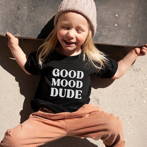 Peut inclure: Un enfant portant un t-shirt noir avec le texte "GOOD MOOD DUDE" en lettres blanches. L'enfant porte un pantalon marron et un bonnet en tricot marron clair.
