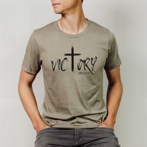 May include: A khaki t-shirt with a black graphic design of a cross and the word "VICTORY" with the text "Song of Songs 2:14" underneath.