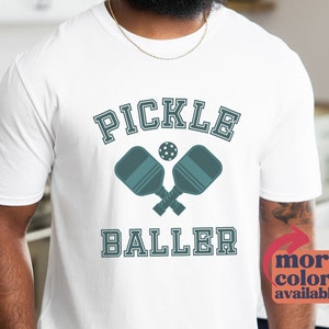 May include: White t-shirt with a blue graphic of two pickleball paddles crossed with a ball in the middle. The text "Pickle Baller" is below the paddles.