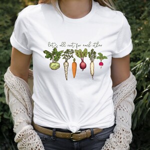 May include: White t-shirt with the text "Let's all root for each other" and a line of illustrated vegetables, including a turnip, carrots, a beet, a radish, and a parsnip.