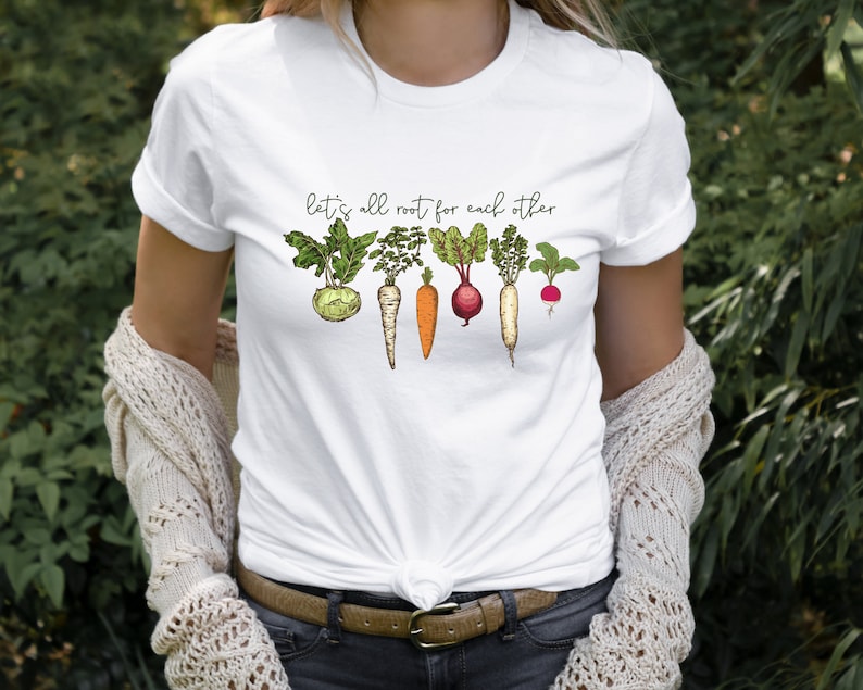 May include: White t-shirt with the text "Let's all root for each other" and a line of illustrated vegetables, including a turnip, carrots, a beet, a radish, and a parsnip.