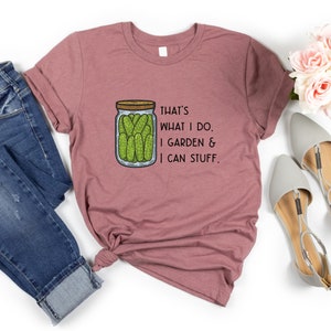 May include: A mauve t-shirt with a graphic of a jar of pickles and the text "That's what I do. I garden & I can stuff."