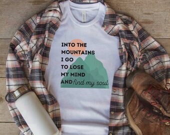 May include: White t-shirt with a graphic design of mountains and a sun. The text reads "Into the Mountains I go to lose my mind and find my soul."