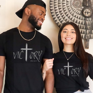 May include: A couple wearing black t-shirts with a silver cross and the word "VICTORY" with the words "John 16:33" underneath.
