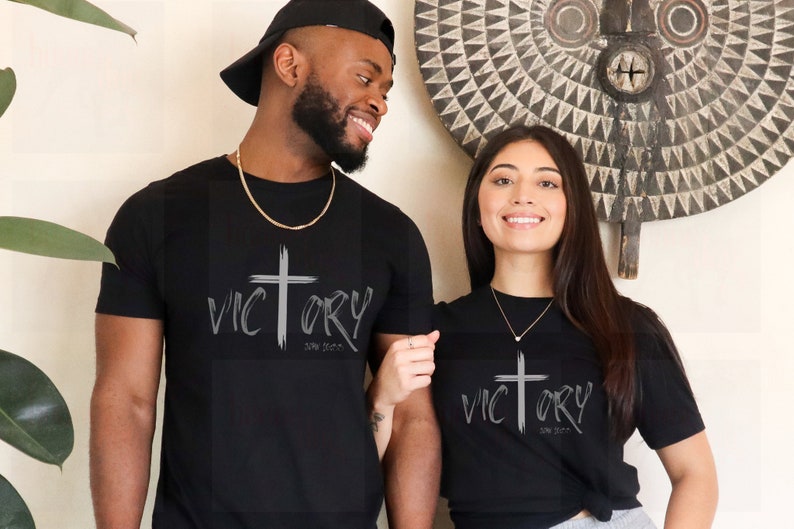 May include: A couple wearing black t-shirts with a silver cross and the word "VICTORY" with the words "John 16:33" underneath.