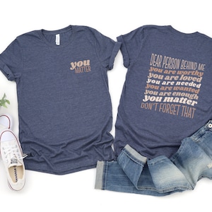 May include: Two navy blue t-shirts with a positive message printed on them. The front shirt says "you matter" in a peachy-pink color. The back shirt says "Dear person behind me you are worthy you are loved you are needed you are wanted you are enough you matter don't forget that" in a peachy-pink color.
