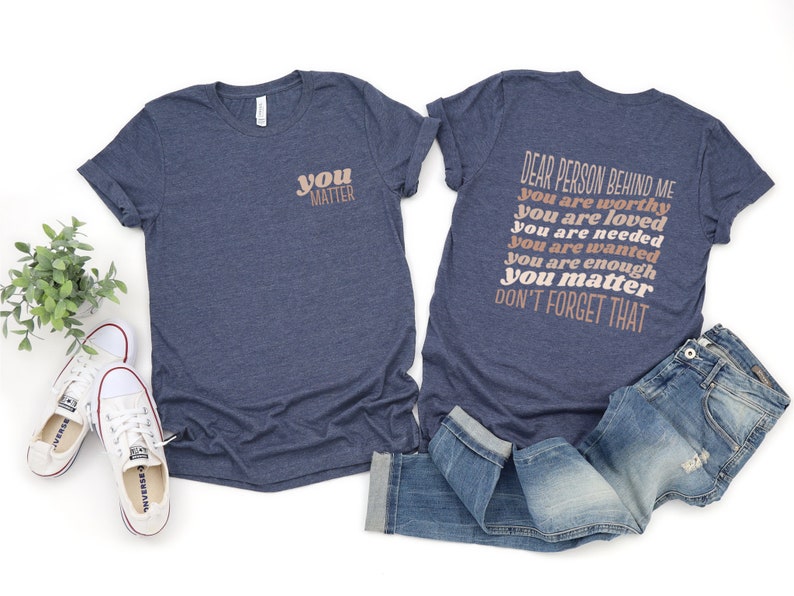 May include: Two navy blue t-shirts with a positive message printed on them. The front shirt says "you matter" in a peachy-pink color. The back shirt says "Dear person behind me you are worthy you are loved you are needed you are wanted you are enough you matter don't forget that" in a peachy-pink color.
