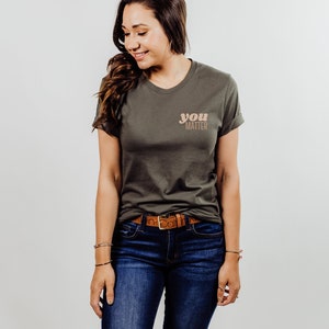 May include: A woman wearing a green t-shirt with the text "you matter" in a peach and brown color scheme. She is wearing blue jeans and a brown belt.