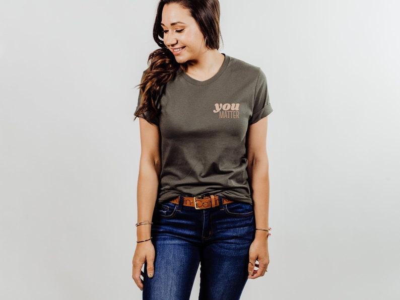 May include: A woman wearing a green t-shirt with the text "you matter" in a peach and brown color scheme. She is wearing blue jeans and a brown belt.