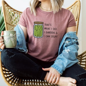 May include: A woman wearing a pink t-shirt with the text "That's what I do. I garden & I can stuff." and a graphic of a jar of pickles. She is sitting in a wicker chair and holding a green mug with a dragonfly design.