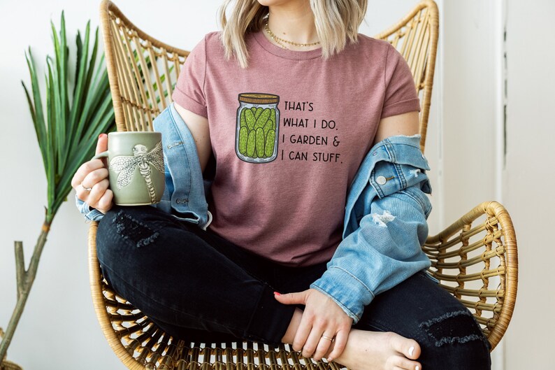May include: A woman wearing a pink t-shirt with the text "That's what I do. I garden & I can stuff." and a graphic of a jar of pickles. She is sitting in a wicker chair and holding a green mug with a dragonfly design.