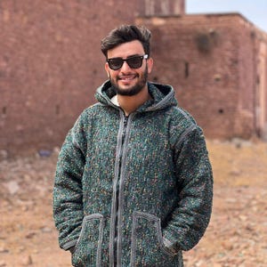 May include: A green hooded jacket with a textured, speckled fabric. The jacket features a full zip, two front pockets, and white trim. The person in the image is wearing sunglasses.