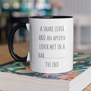 Peut inclure: Une tasse à café blanche avec une anse noire et un bord noir. La tasse porte une inscription noire qui dit "A snake lover and an Apteryx lover met in a bar.  The end."