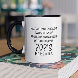 May include: White coffee mug with a black handle and black rim. The mug has black text that reads "Half a cup of sarcasm, two spoons of profanity and a pinch of truth equals Pop's Persona". The mug is sitting on a stack of books.