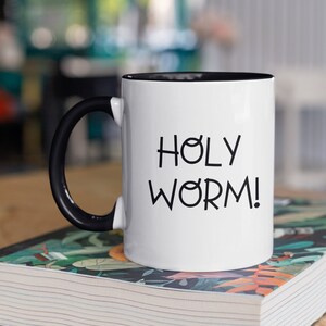 May include: White coffee mug with a black handle and black rim. The mug has the text "HOLY WORM!" printed in black on the front.