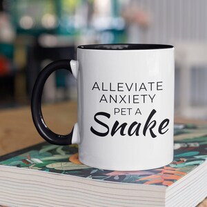 Peut inclure: Une tasse à café blanche avec une anse noire et un bord noir. La tasse porte une inscription noire qui dit "Alleviate Anxiety Pet a Snake". La tasse est posée sur un livre avec un motif floral coloré.
