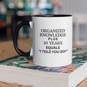 Peut inclure: Une tasse à café blanche avec une anse noire et un bord noir. La tasse porte une inscription noire qui dit "Organized Knowledge Plus 10 Years Equals "I Told You So!" La tasse est posée sur une pile de livres à motifs floraux.