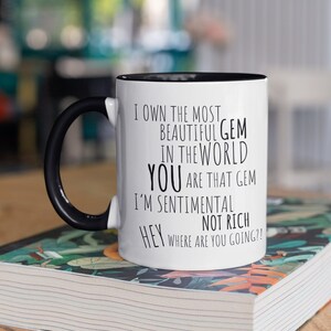 May include: White coffee mug with a black handle and black rim. The mug has black text that reads "I own the most beautiful gem in the world you are that gem I'm sentimental not rich hey where are you going?!"