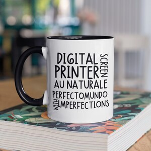 May include: White ceramic mug with a black rim and handle. The mug has black text that reads "DIGITAL SCREEN PRINTER AU NATUREL PERFECTO MUNDO WITH IMPERFECTIONS". The mug is sitting on a book with a floral pattern.