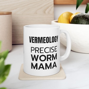 May include: White ceramic mug with the words "VERMEOLOGY PRECISE WORM MAMA" printed in bold black letters. The mug is on a beige coaster. The background includes a wooden cutting board, a bowl with fruit, and greenery.
