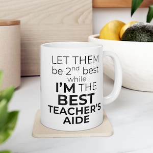 Könnte beinhalten: Weiße Keramik-Tasse mit dem Text "LET THEM be 2nd best while I'M THE BEST TEACHER'S AIDE" in Schwarz. Die Tasse steht auf einem beigen Untersetzer, mit verschwommenem Hintergrund.