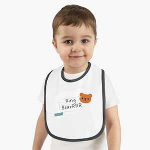 May include: White baby bib with a dark gray trim. The bib features the text "Ring BearRRR" and a cartoon bear illustration. The bib is designed for infants and toddlers.