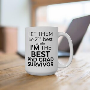 May include: White ceramic mug with the text "LET THEM be 2nd best while I'M THE BEST PhD GRAD SURVIVOR" in black. The mug has a large handle and is sitting on a wooden surface. A laptop is visible in the background.