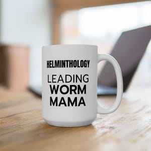 May include: White ceramic mug with the text "HELMINTHOLOGY LEADING WORM MAMA" in black. The mug is on a wooden surface, with a laptop in the background. The mug has a large handle.