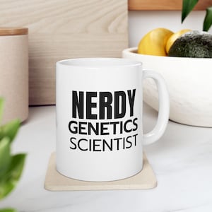 Op de afbeelding: Witte keramische mok met de tekst "NERDY GENETICS SCIENTIST" in zwart. De mok staat op een beige onderzetter. De achtergrond is wazig, met een houten oppervlak en een fruitschaal.