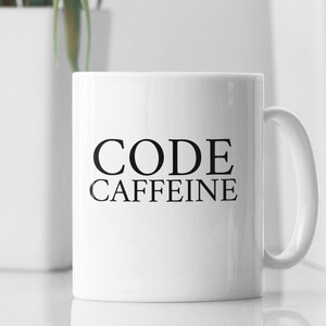 May include: White ceramic mug with the words "CODE CAFFEINE" printed in black. The mug has a curved handle and is set against a blurred background with a white planter and a stack of books.