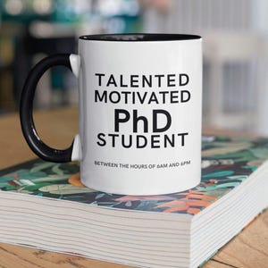 May include: White ceramic mug with a black rim and handle. The mug has black text that reads "Talented Motivated PhD Student Between the hours of 6AM and 6PM".