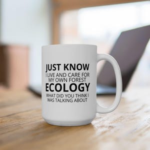 May include: A white ceramic mug with a black handle. The mug has the text "JUST KNOW I LIVE AND CARE FOR MY OWN FOREST ECOLOGY WHAT DID YOU THINK I WAS TALKING ABOUT" printed on the front. The mug is on a wooden surface.