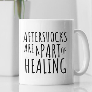May include: White ceramic mug with black text that reads "Aftershocks are a part of healing".